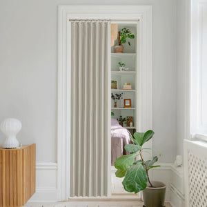 Light beige pleated fabric curtain panel hanging on a silver rod in a doorway, partially revealing shelves and plants.
