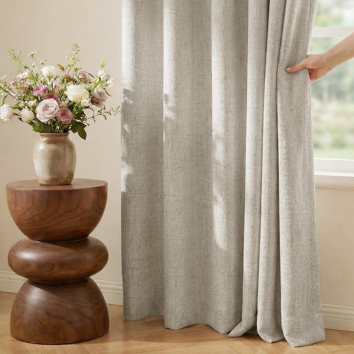 Textured light beige curtain panel drapes by a window. Hand pulls back the woven fabric, revealing light.