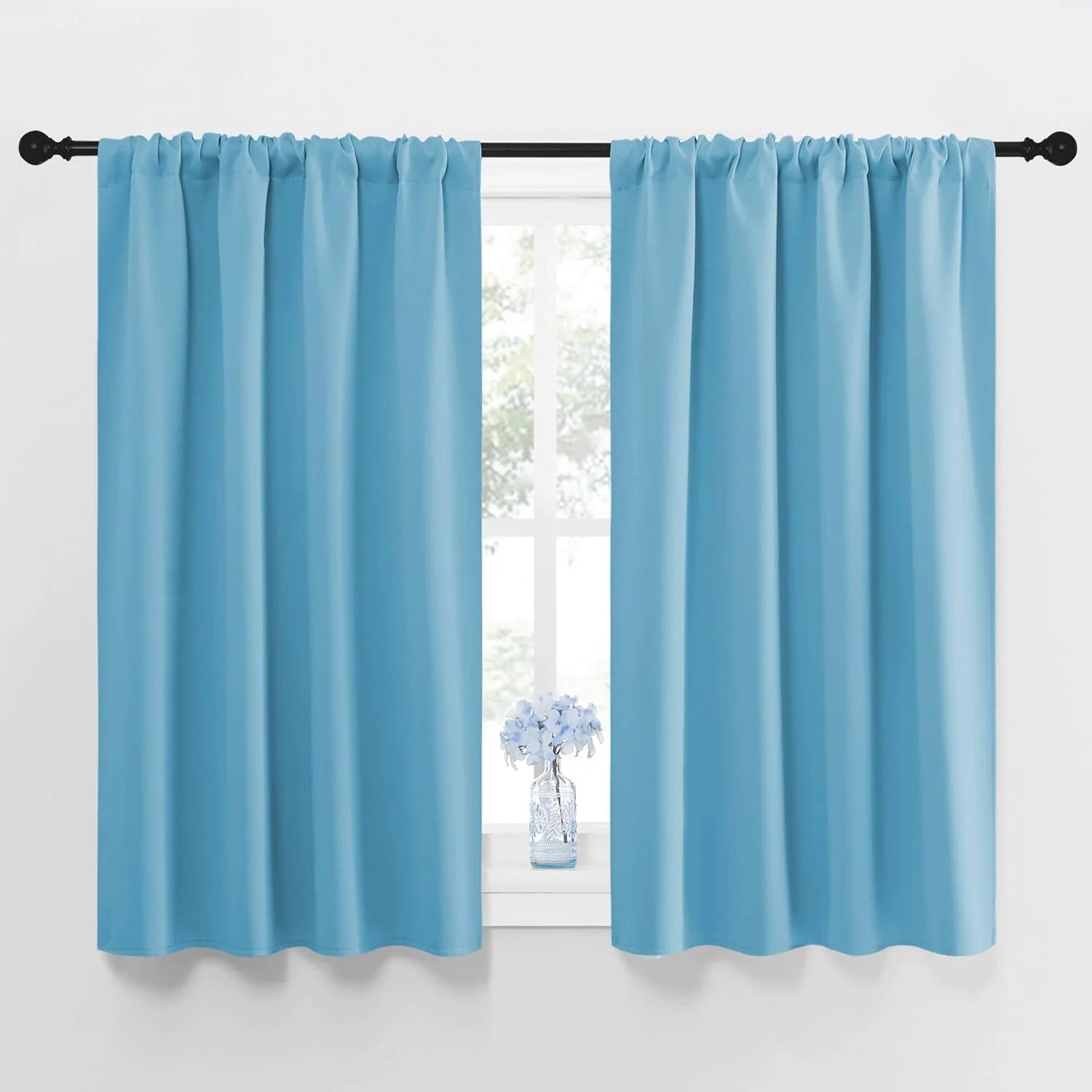 Two light blue rod pocket short curtains hung on a black rod over a window with a vase