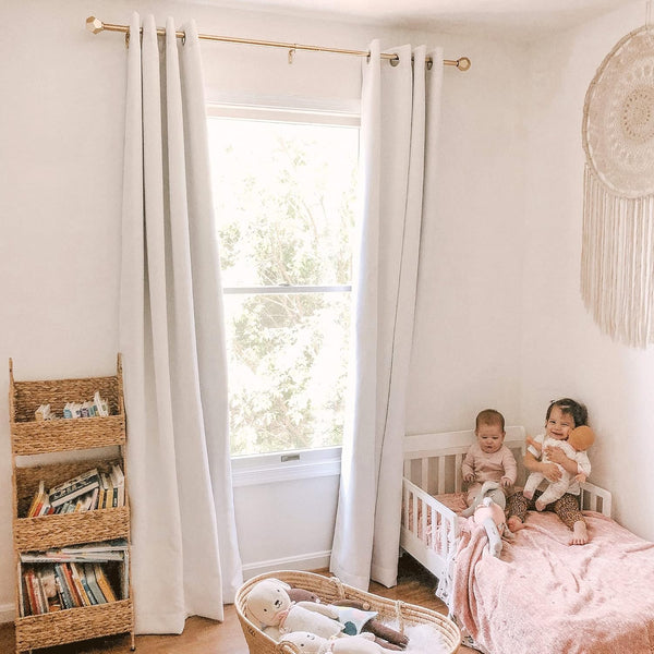 Light grey grommet blackout curtains hanging on a gold rod in a bright nursery with white walls.