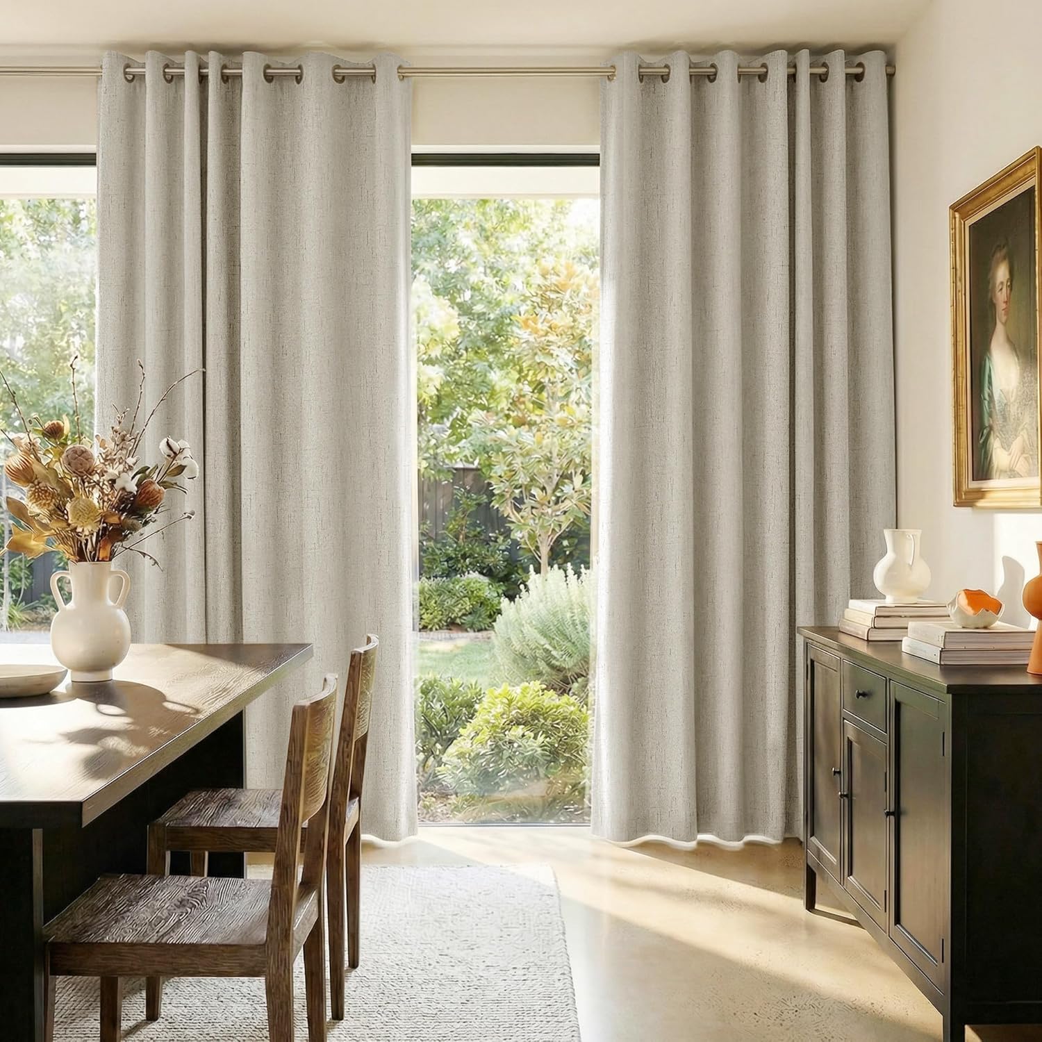 Textured light grey grommet top curtains frame large windows overlooking a bright garden in a dining room.