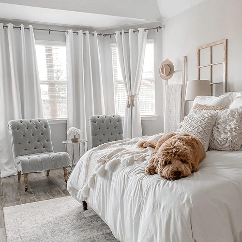 Bright bedroom decor featuring two light grey tufted accent chairs, white bedding, and grommet curtains.
