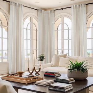 Light natural linen pleated curtains hang on a dark rod, framing tall arched windows in a bright modern living room.
