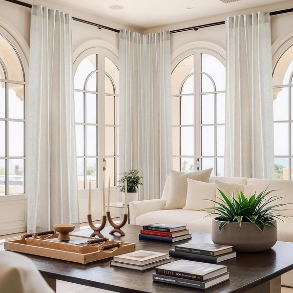 Light natural linen pleated curtains hang on a dark rod, framing tall arched windows in a bright modern living room.