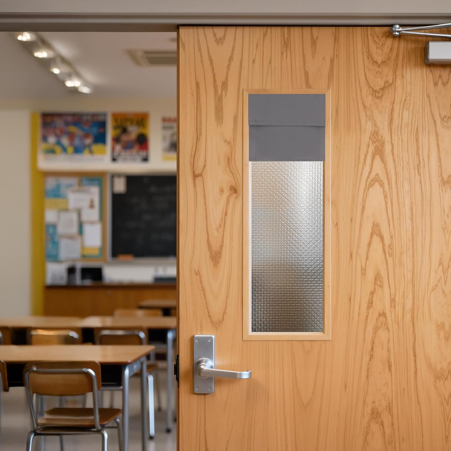 Light wood door with frosted glass window and grey fabric blind, ajar to a classroom.
