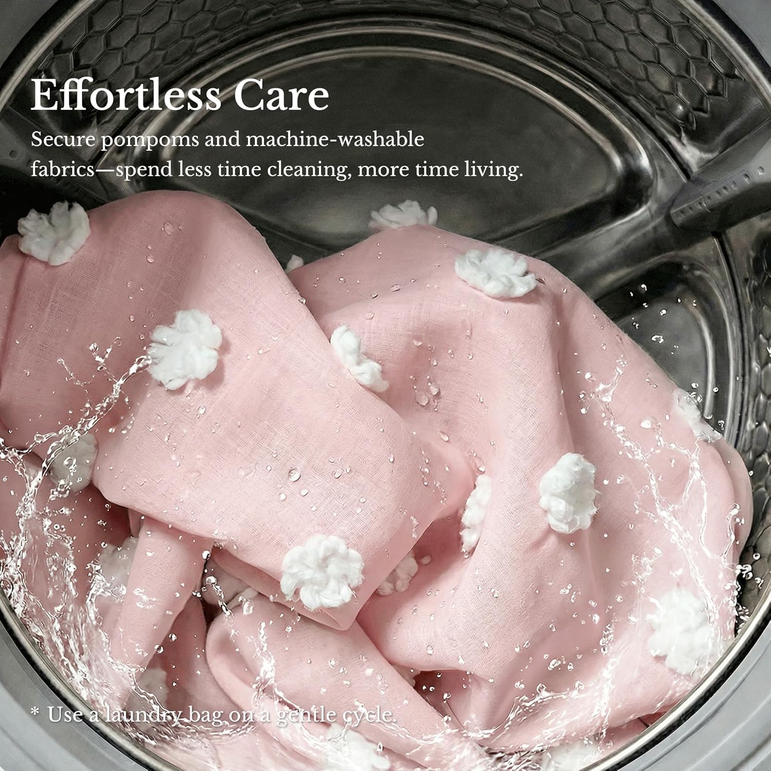 Close-up of pink fabric with white pom-poms washing in a metallic machine drum, water splashing.
