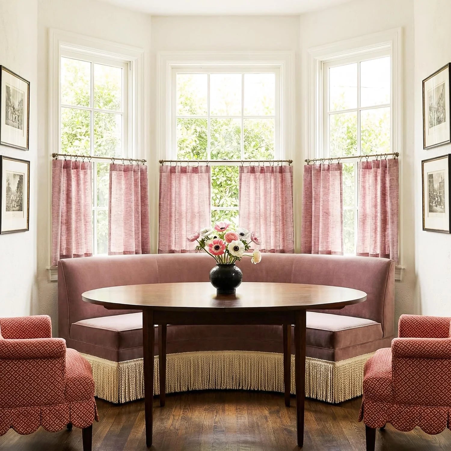Elegant mauve banquette with fringe skirt in a bay window dining nook, round wood table, and red patterned armchairs.