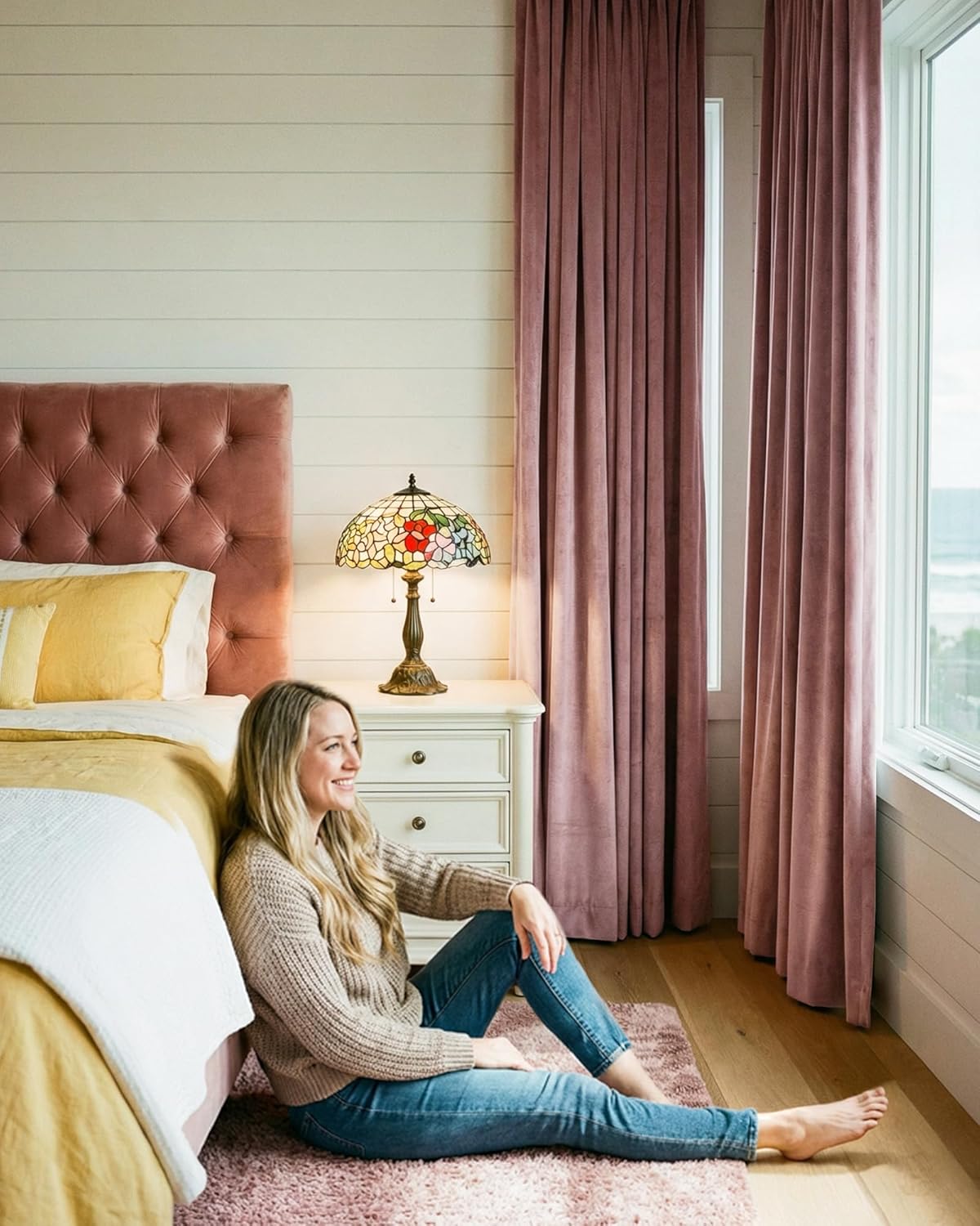 Mauve velvet pleated curtains frame an ocean view window next to a matching tufted headboard in a cozy bedroom.