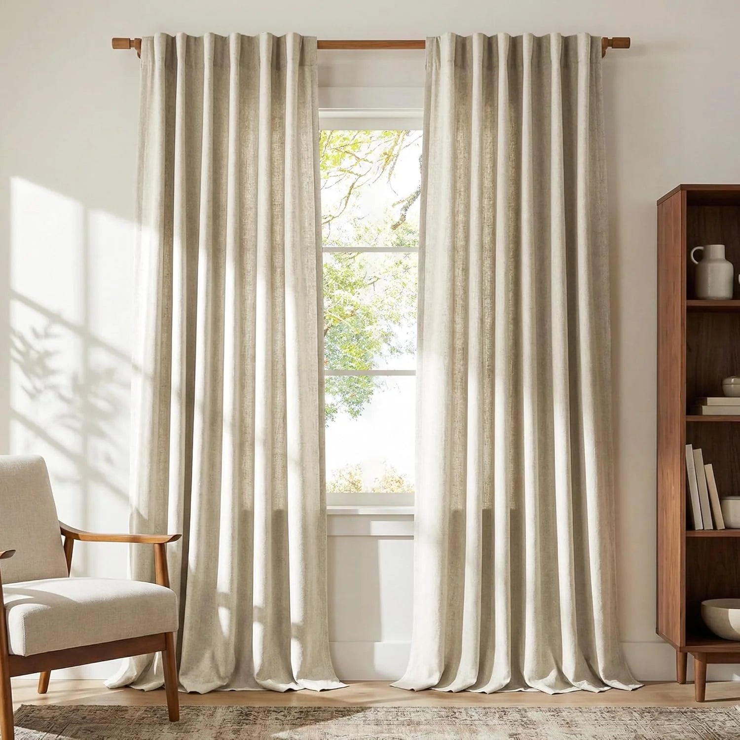 Natural linen-look curtains hanging on a wooden rod in a bright living room with a cream chair and bookshelf.