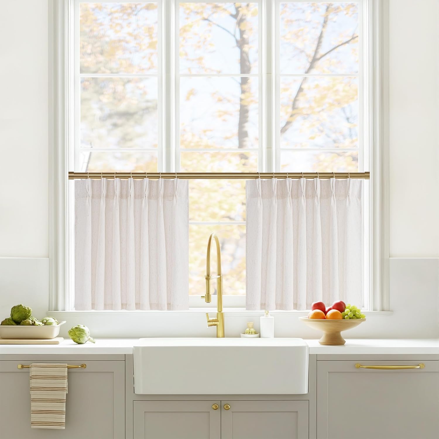 Natural linen semi-sheer cafe curtains with pleated tops on a brass rod over a kitchen sink.