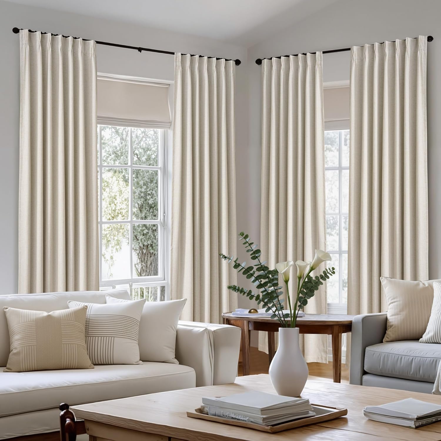 Light beige linen-textured pinch pleat drapes on black rods, framing sunny living room windows.