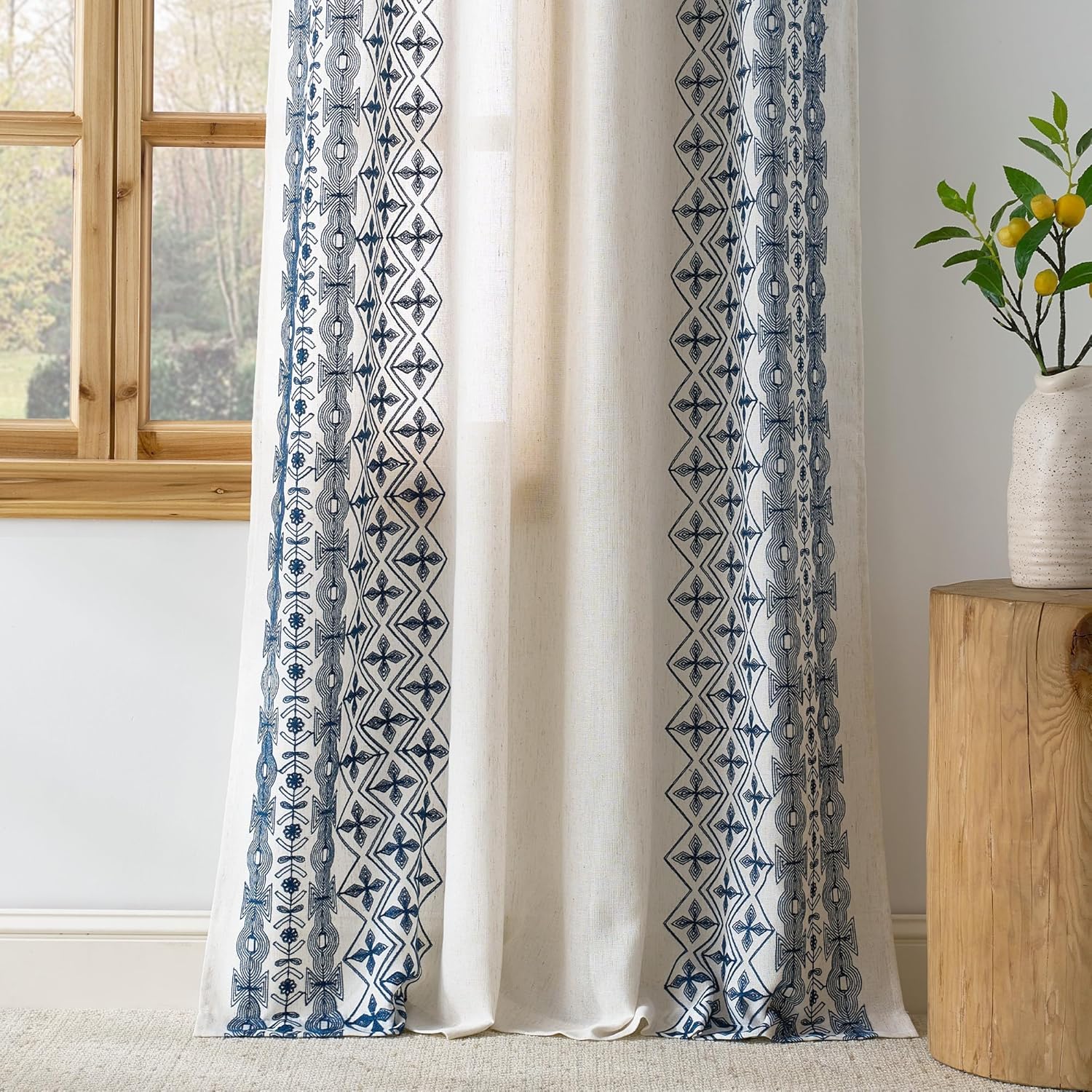 Off-white linen curtain features vertical blue geometric embroidery, hanging by a wooden window.