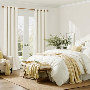 Off-white grommet top curtains frame French doors in a bright bedroom with cozy white bedding and yellow throw.