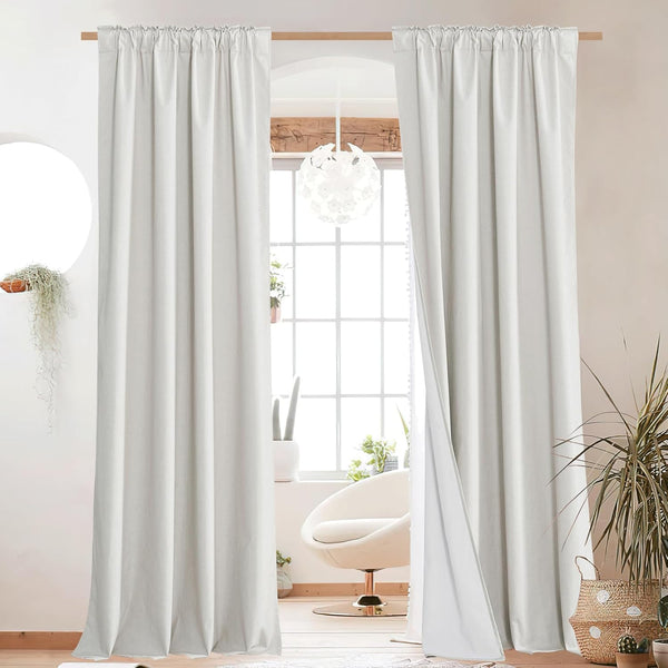Off-white rod pocket blackout curtains with thick fabric hanging on a wooden rod in a bright room.