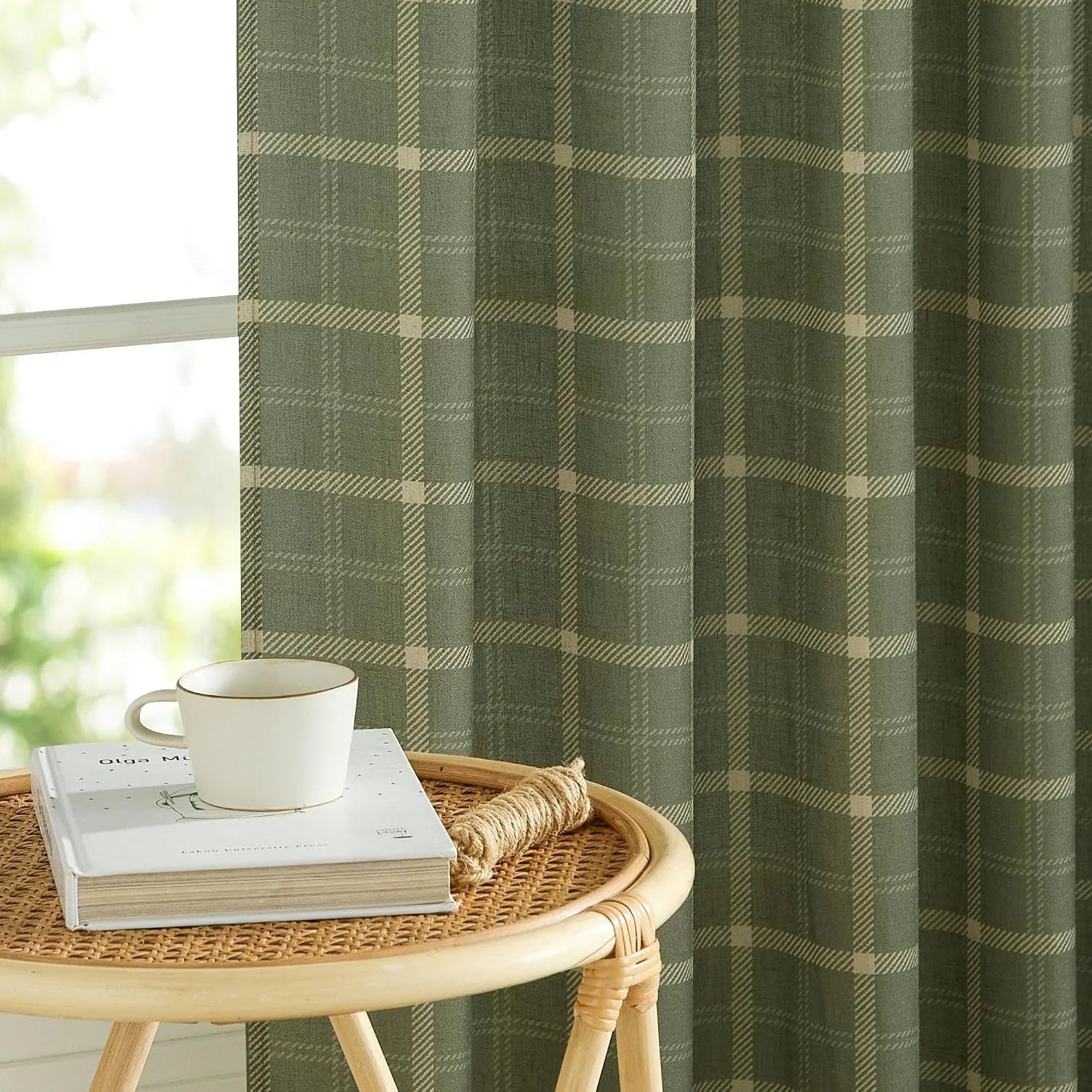 Olive green plaid curtain beside a window with a rattan side table, book and cup