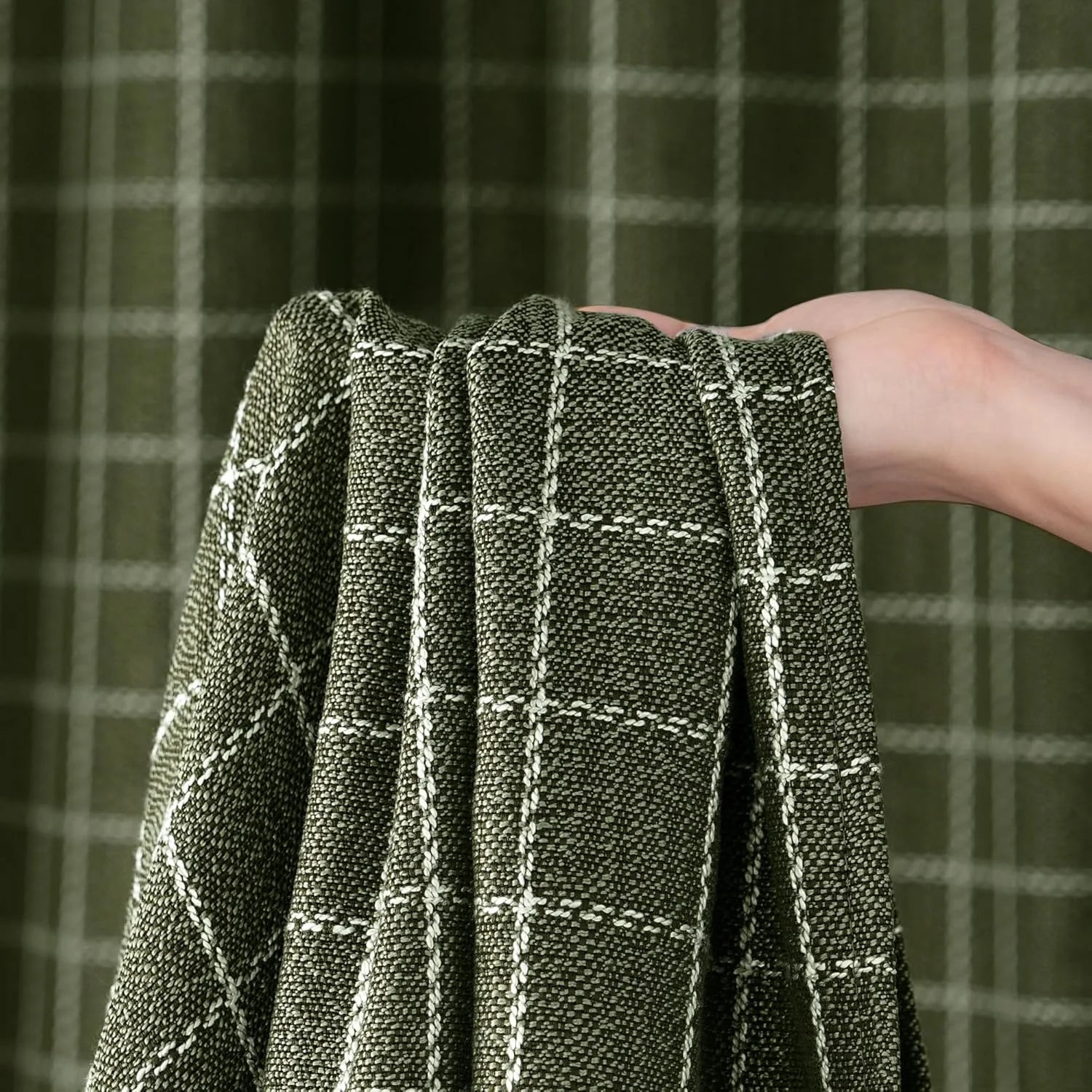 A hand holds a sample of olive green curtain fabric with a white windowpane plaid pattern.