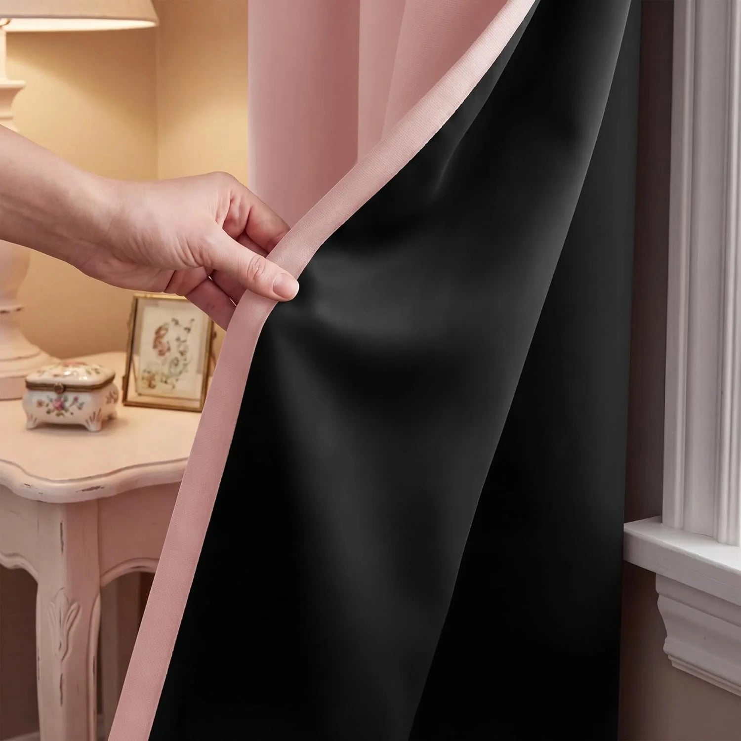 Hand pulling back a pink curtain to reveal its opaque black blackout lining in a warm room