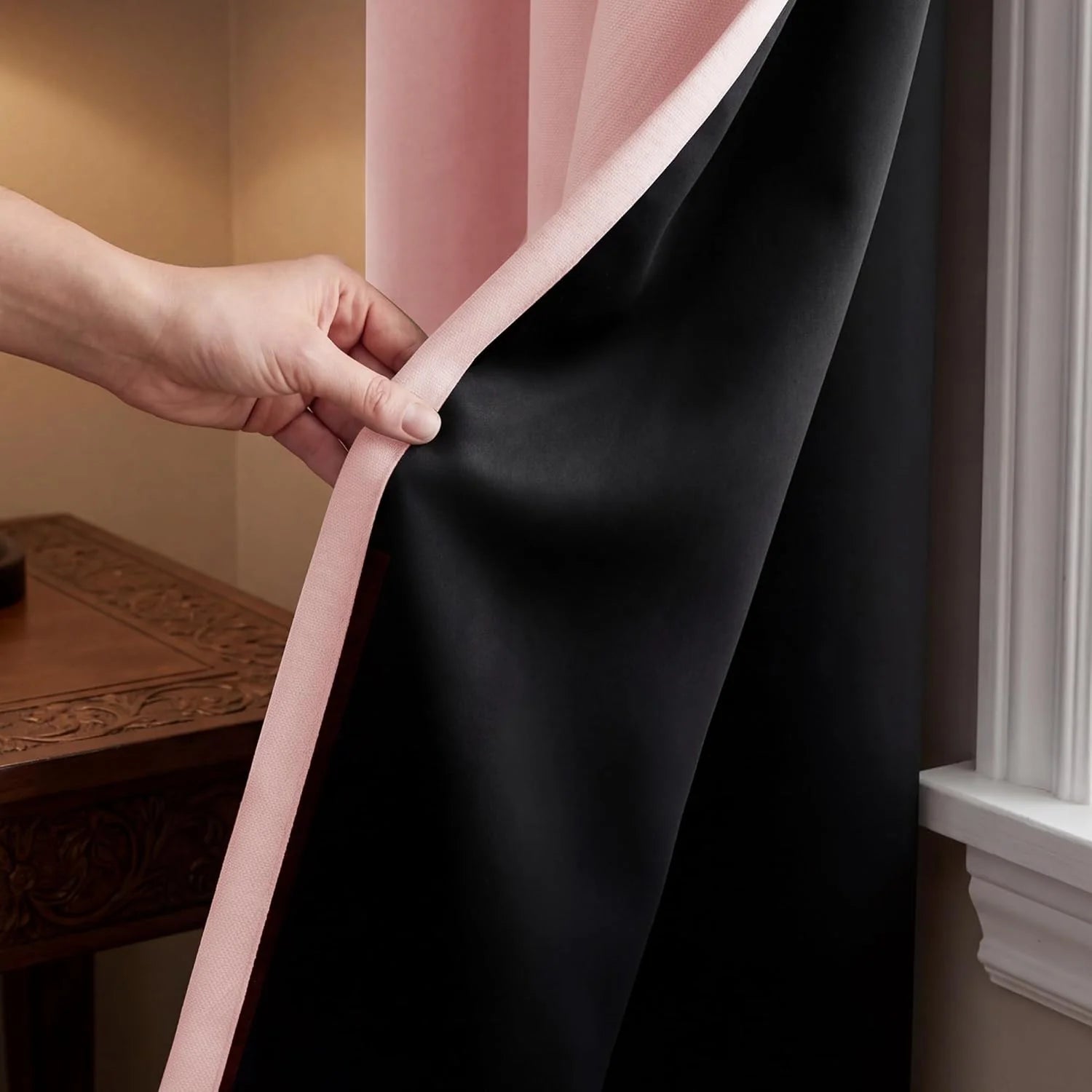 A hand pulls back a pink curtain to reveal its black blackout lining, with a carved wooden table in the background.