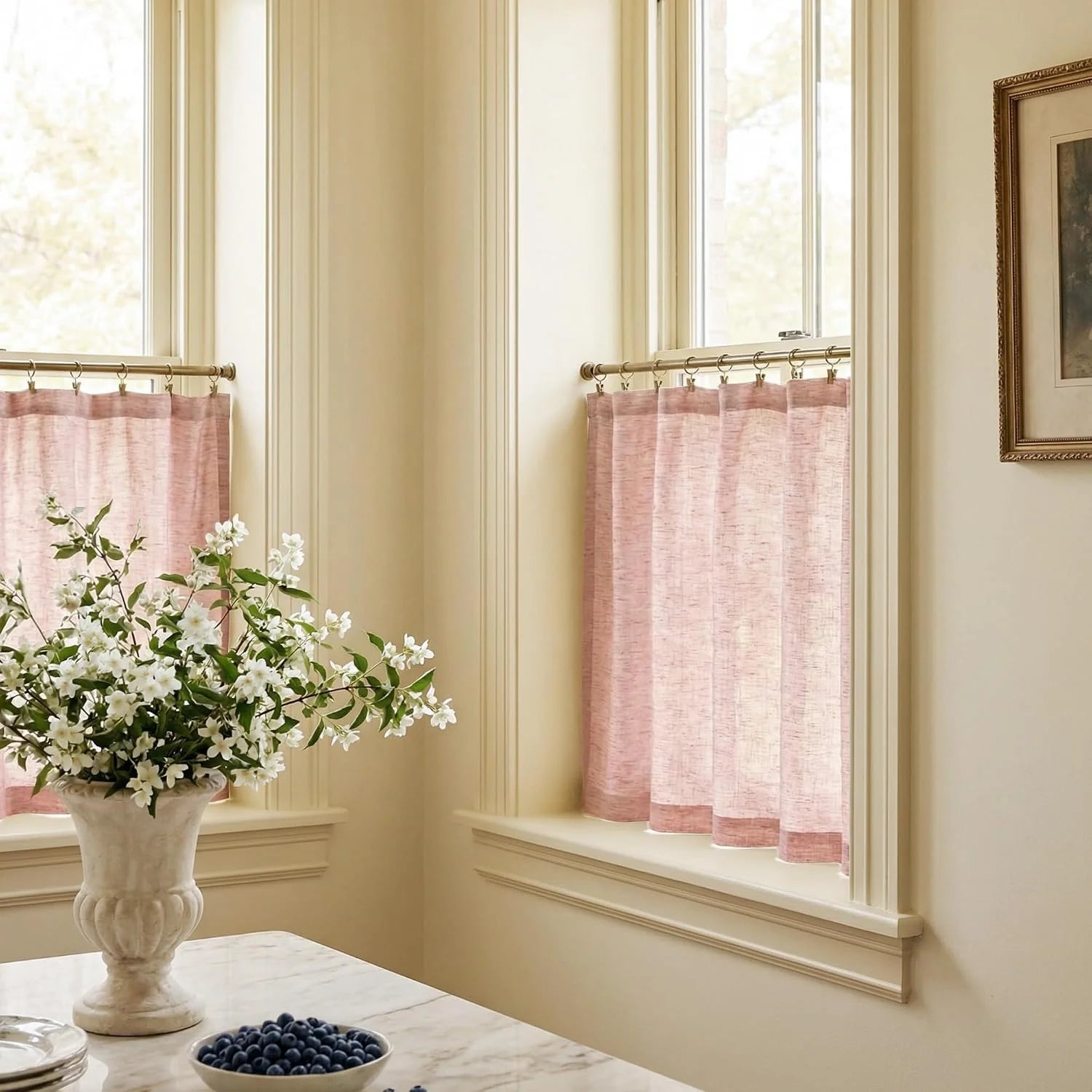 Two windows with sheer pink cafe curtains hung on brass rods in a bright kitchen or dining area. A marble table holds white flowers and blueberries.