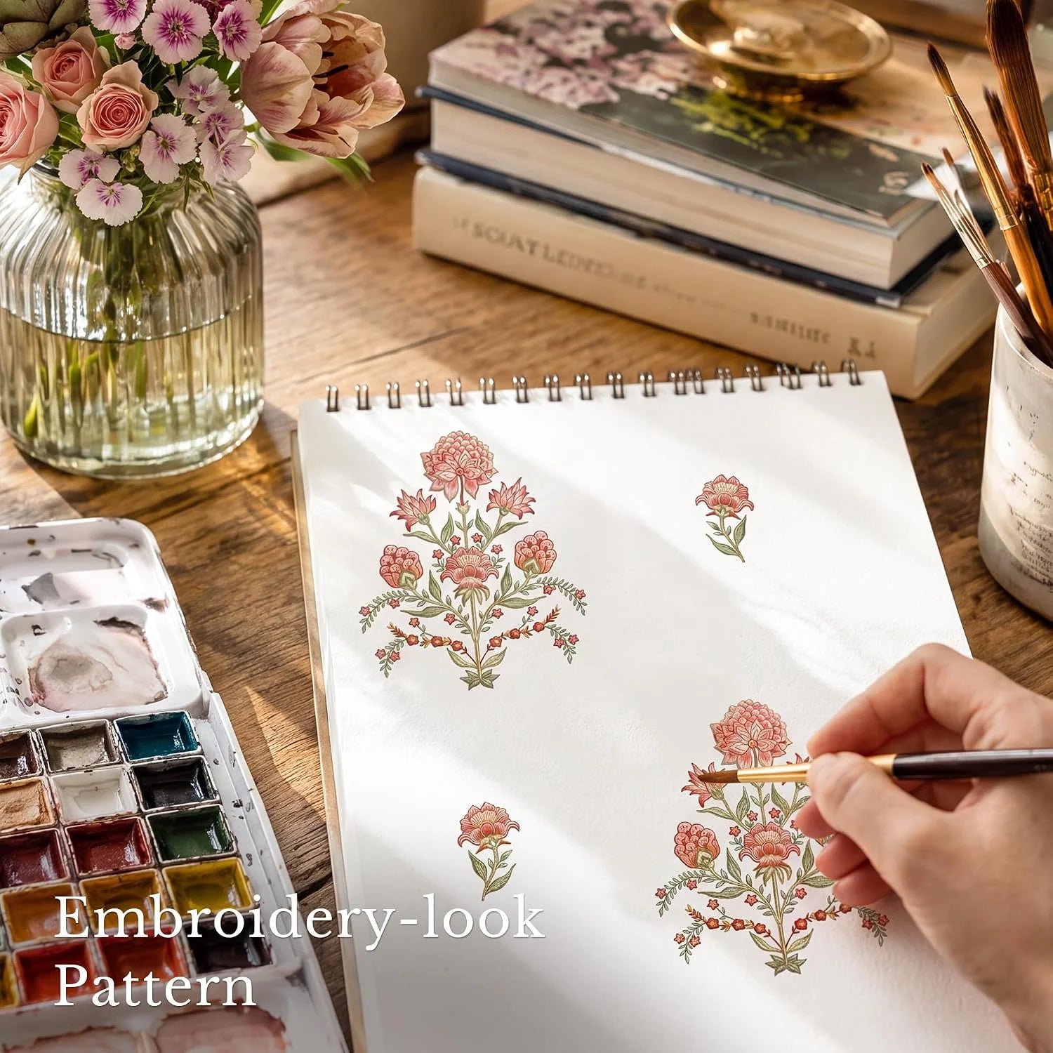 Hand painting a pink floral embroidery-look pattern in a sketchbook on wooden desk