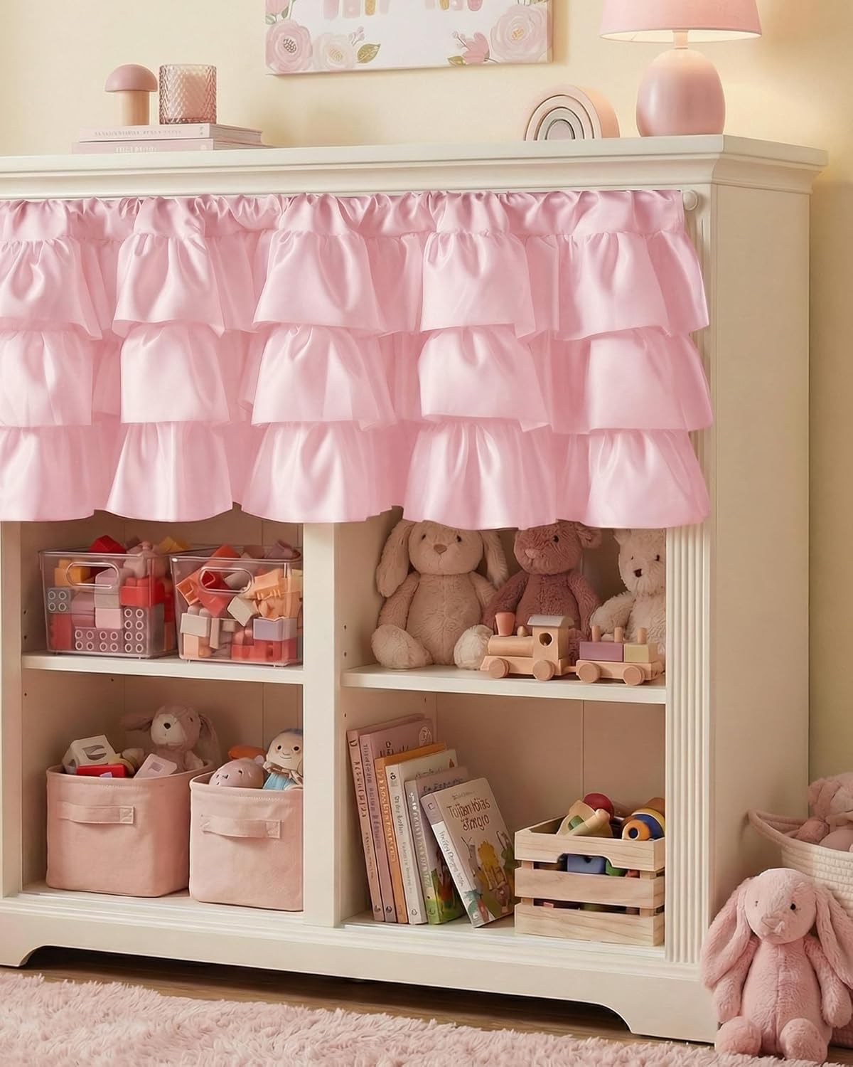 Soft pink tiered ruffled valance hanging over white bookcase shelves in a cozy nursery or child's bedroom.