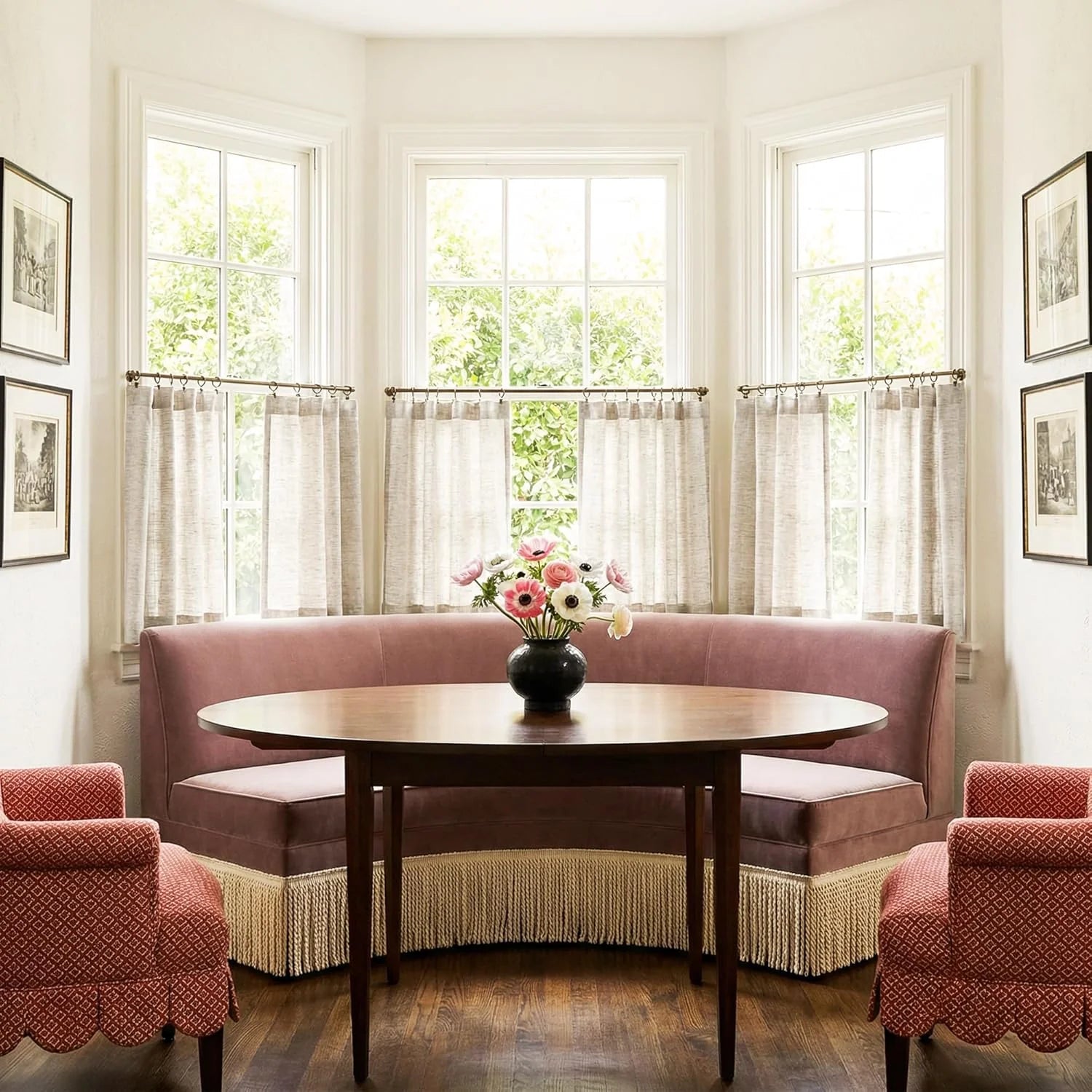 Elegant pink velvet banquette, round wood table, red chairs, and cafe curtains in dining nook.