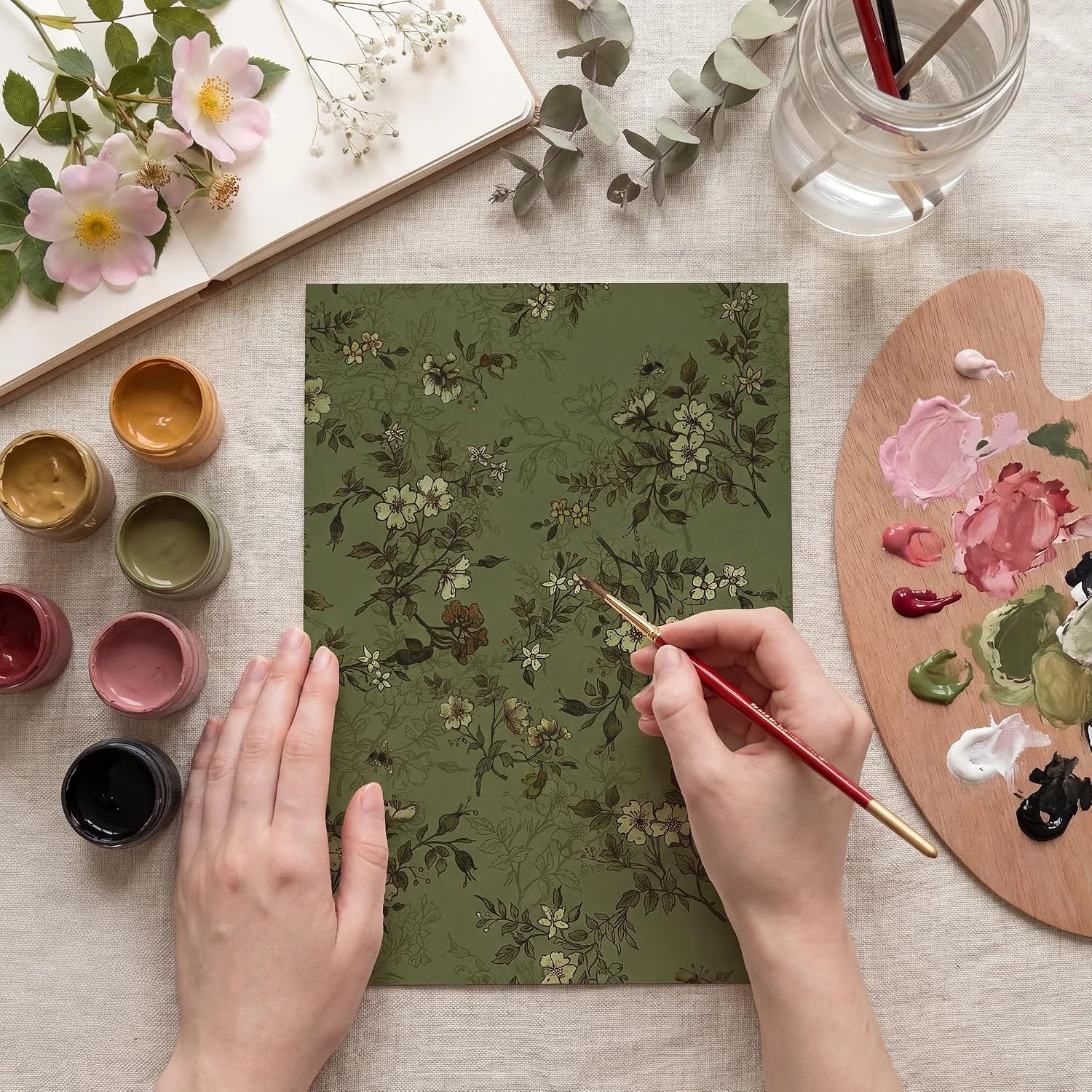 Hands painting a sage green floral sheet with brushes, palette and flowers