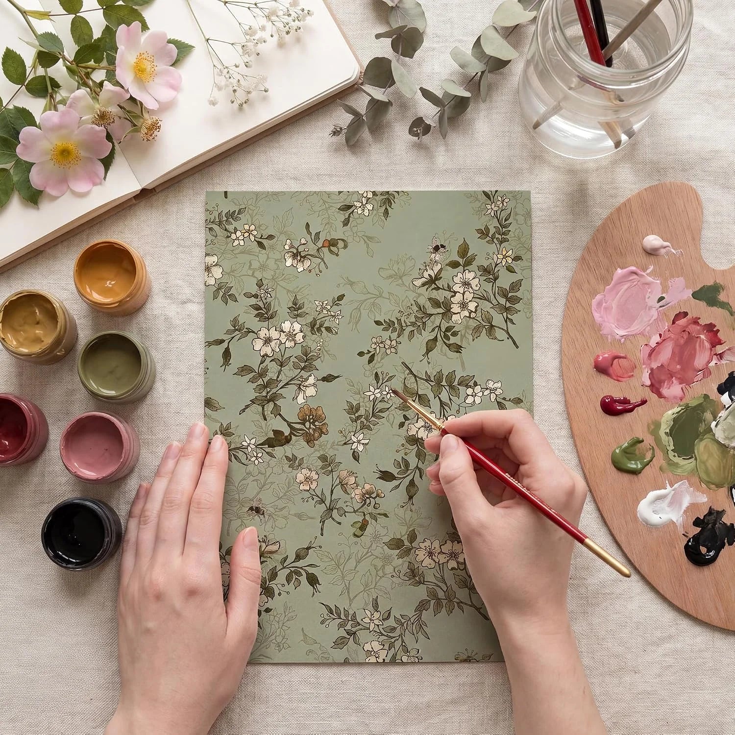 Hands painting a sage green floral paper design with brushes, palette, and flowers