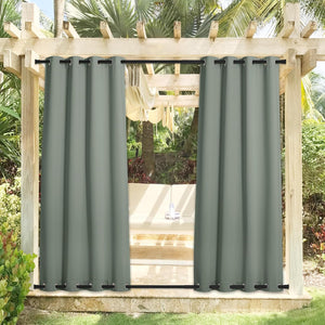 Pair of sage green outdoor grommet top curtains on a pergola patio surrounded by tropical plants.