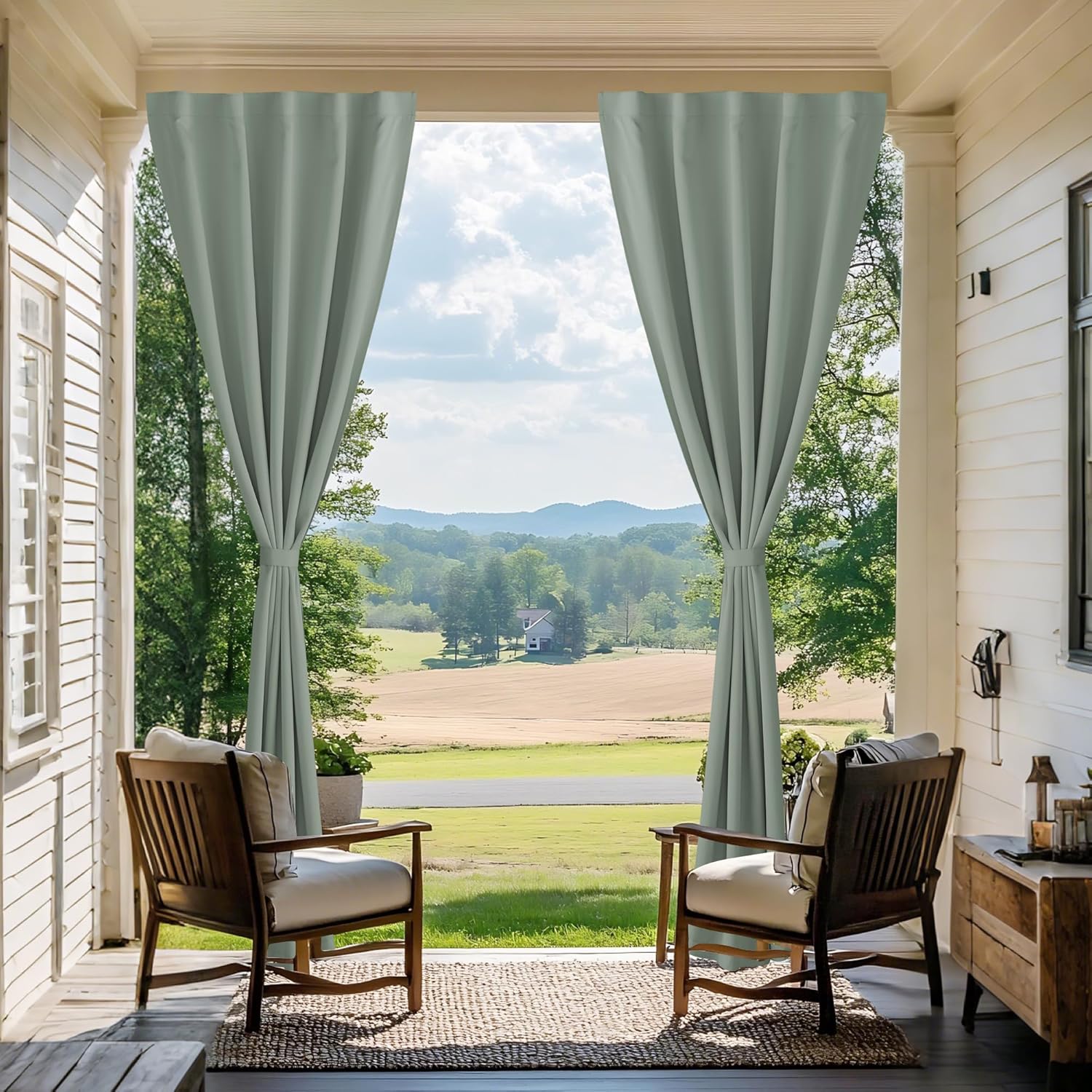 Pair of light sage green outdoor patio curtains with tie-backs on a white porch, framing a countryside view.