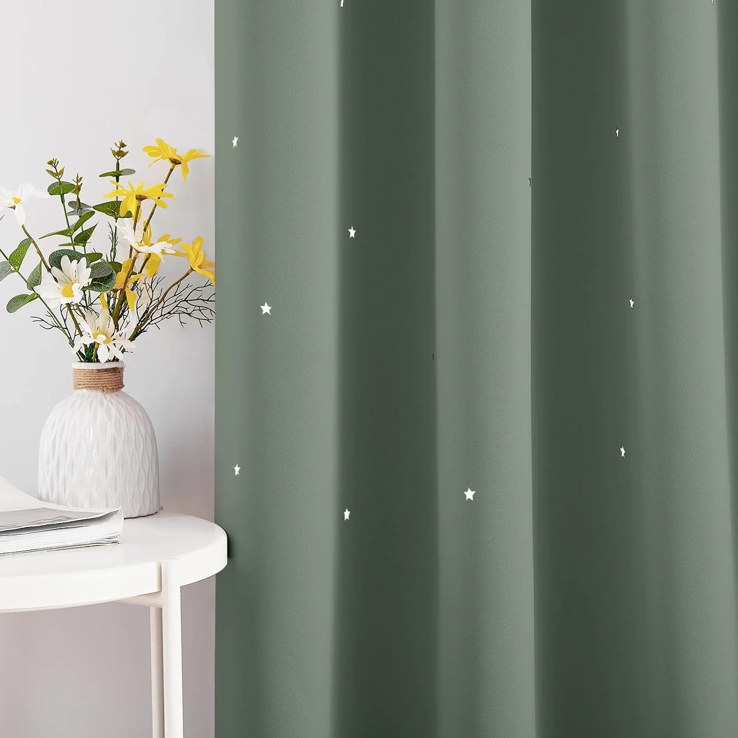 Sage green curtain with small white star pattern beside a white side table and vase