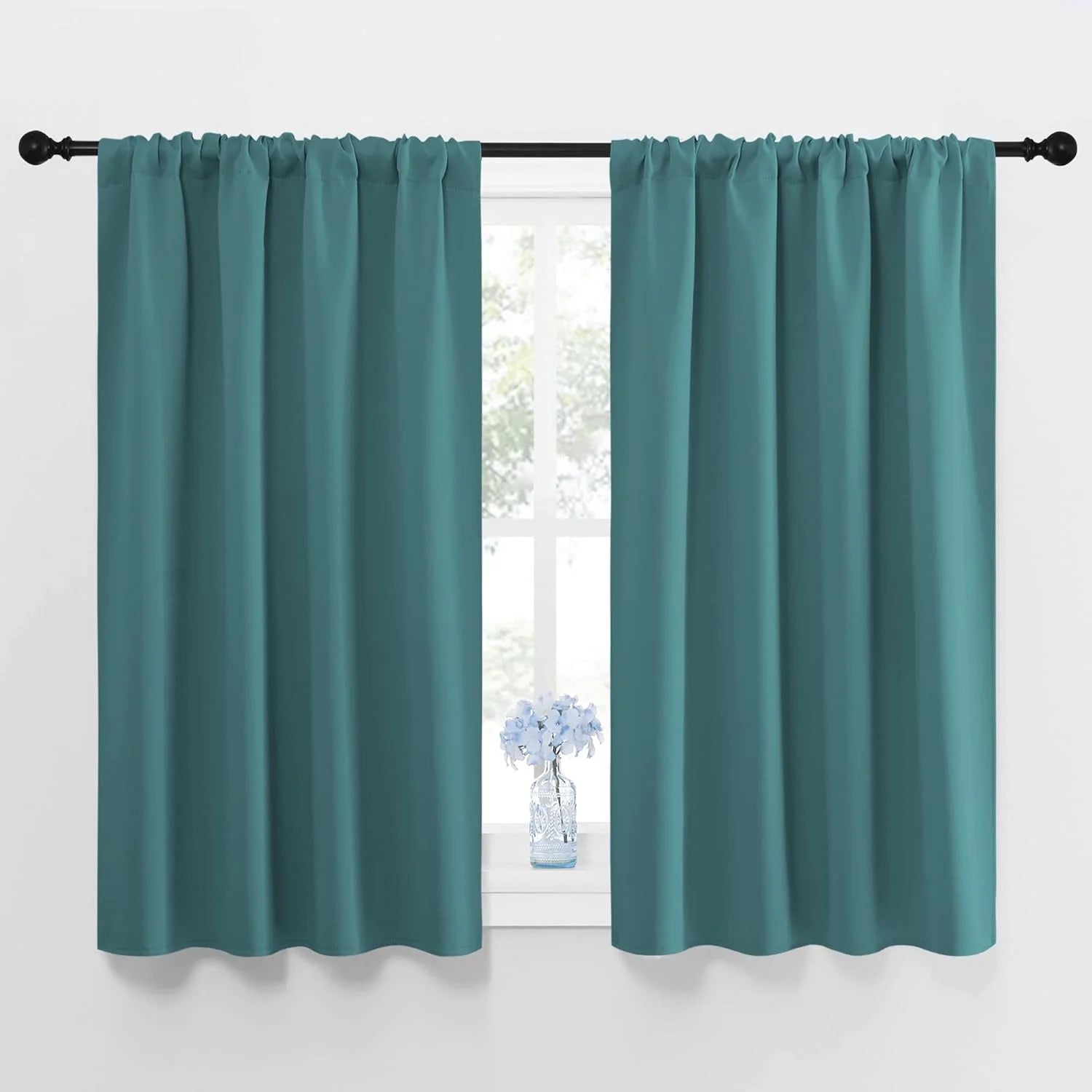 Pair of short teal rod pocket curtains on a black rod, over a window with blue flowers on the sill.
