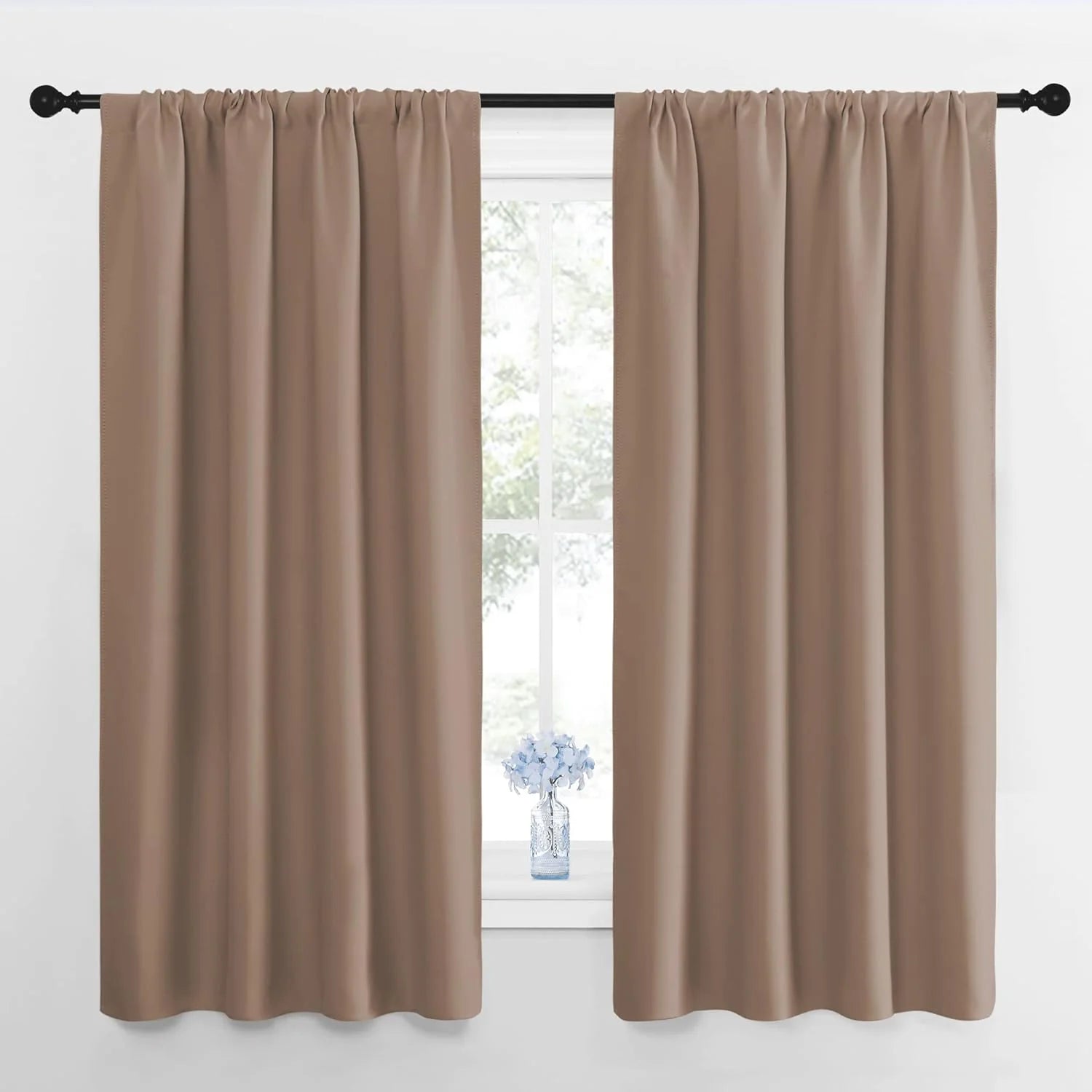 Pair of taupe rod pocket blackout curtains on a black rod over a window with a small vase of blue flowers.
