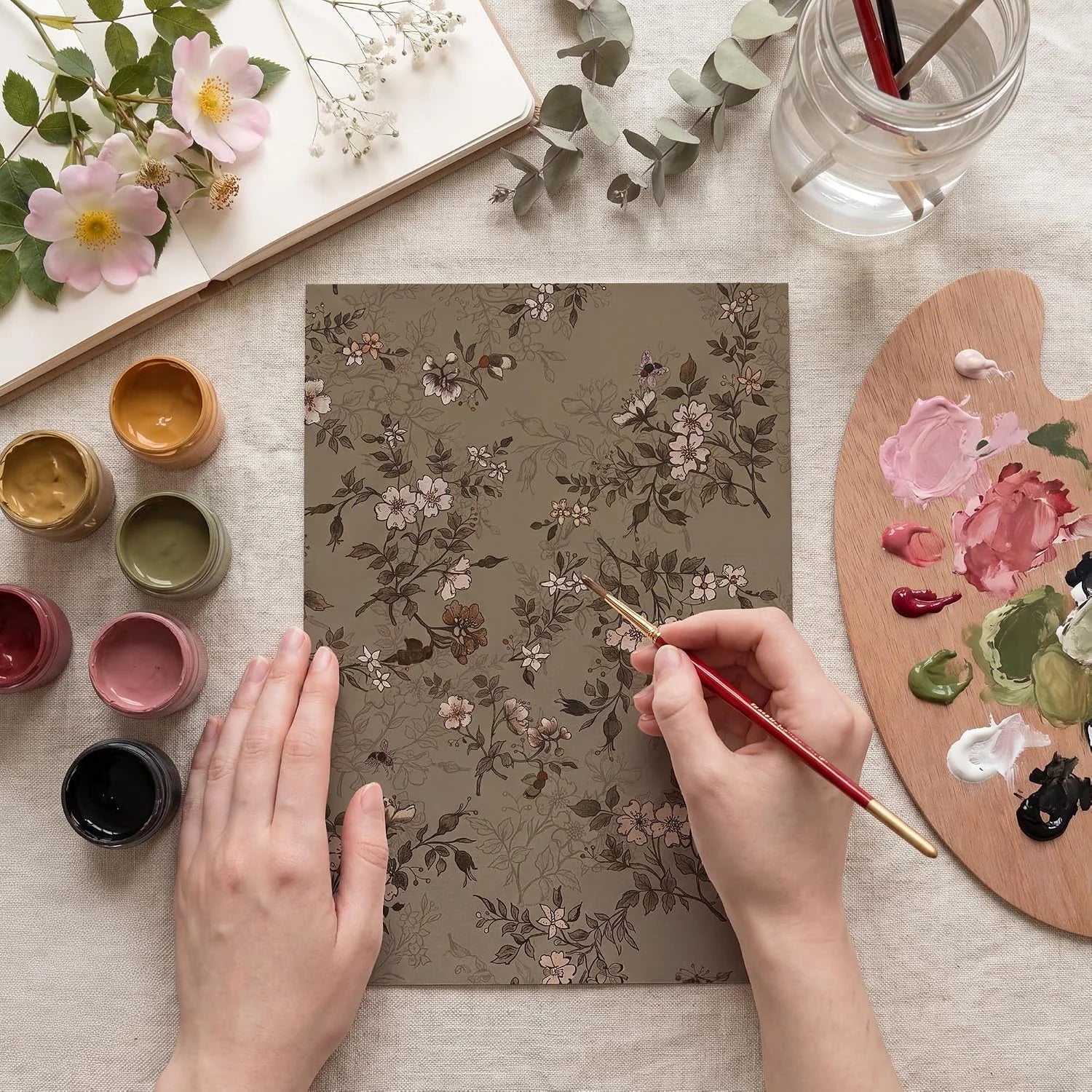 Hands painting a taupe floral art sheet with brushes, palette, and soft pink flowers