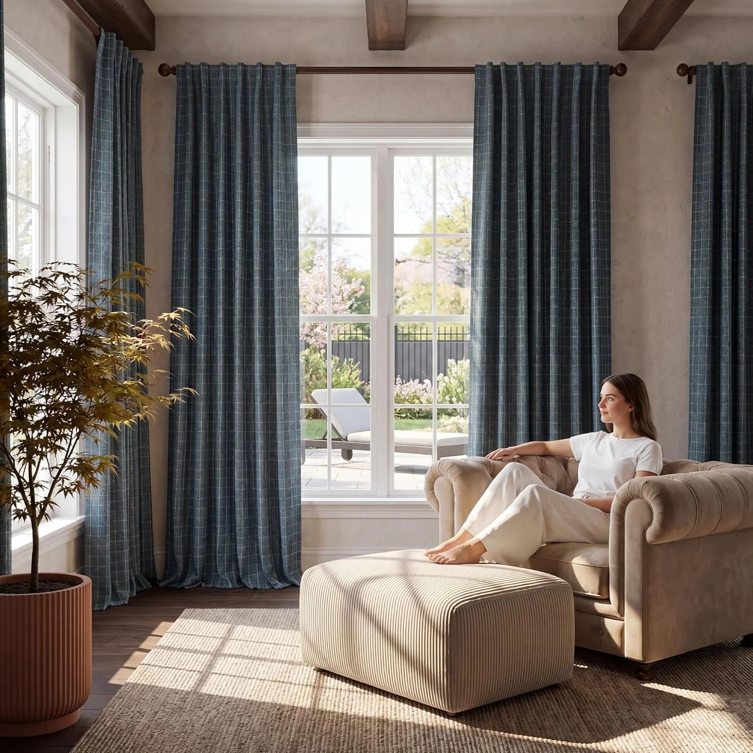 Woman relaxing in beige armchair with ottoman in sunny living room with patterned teal curtains.