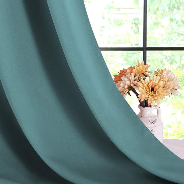 Close-up of a smooth teal solid blackout curtain panel draped beside a window with a vase of flowers.