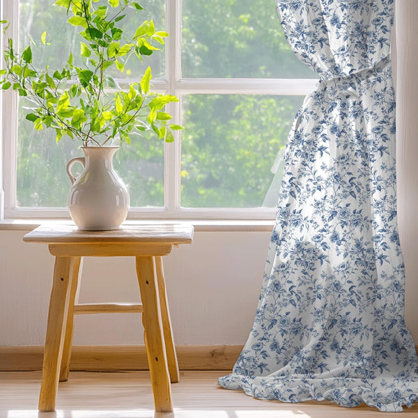 White curtain with blue floral rose pattern and ruffled bottom edge, tied back from a sunlit window.