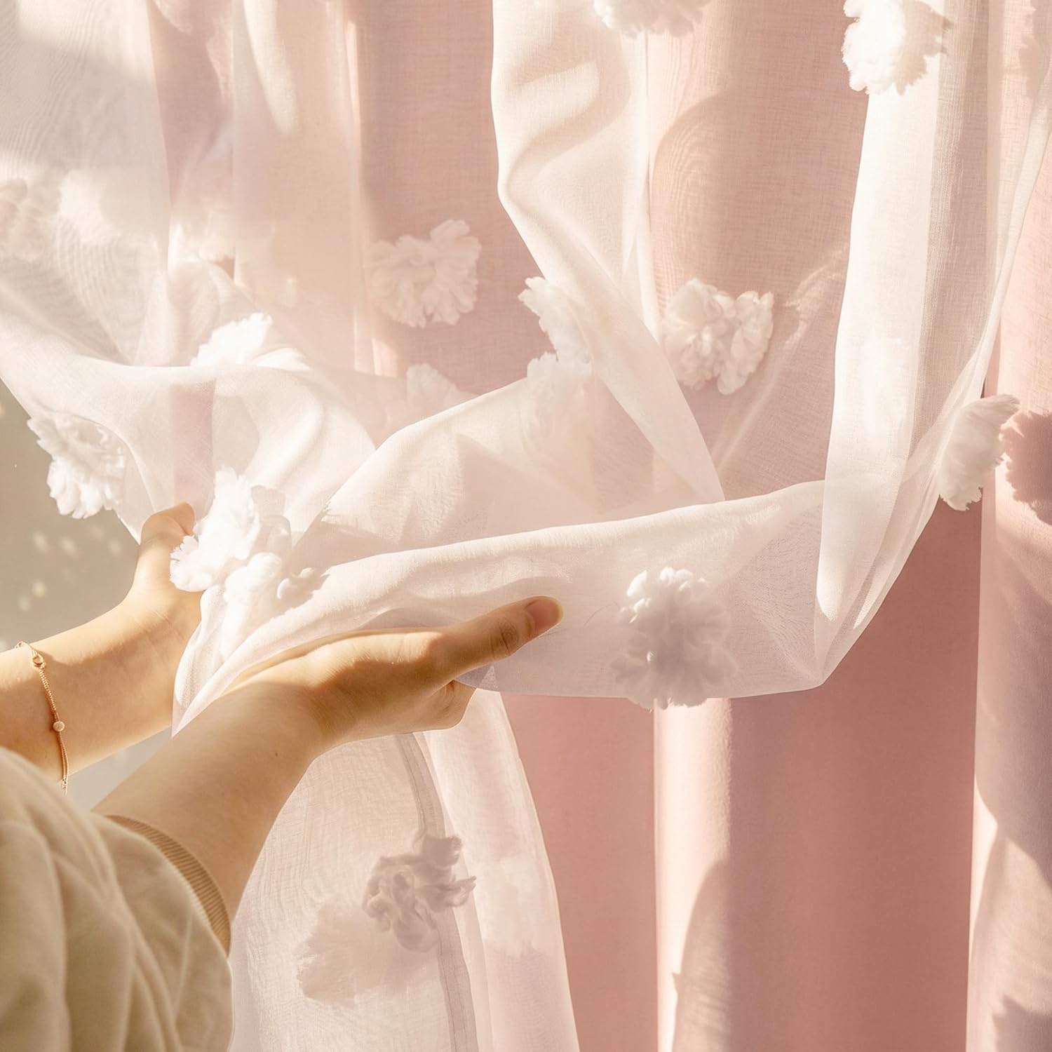 Delicate white sheer voile curtain with fluffy pom-pom flower details, held by hands, sunlight filtering.