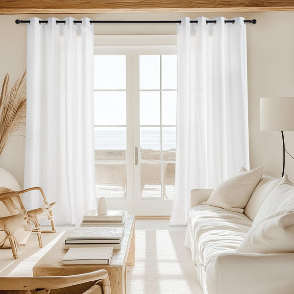 White semi-sheer grommet top curtains hanging on a black rod in a bright living room overlooking the ocean.