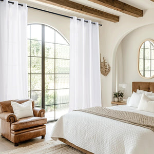 White grommet top sheer curtains hanging on a black rod, framing a large arched window in a bright bedroom.