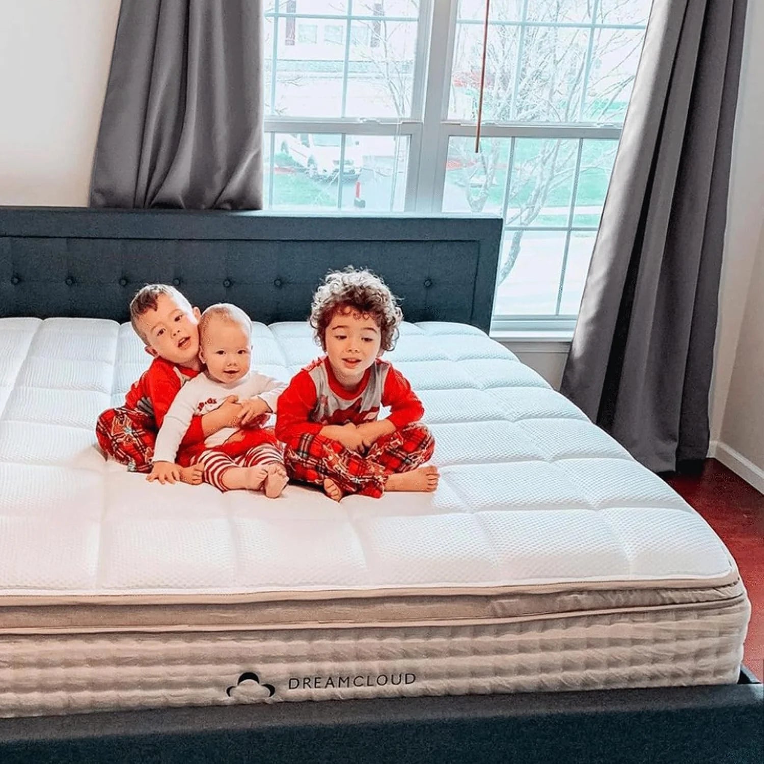 Three children in red pajamas sitting on a white quilted mattress in a bright bedroom