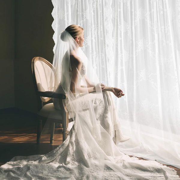 Elegant bride in white lace wedding dress with long train and sheer veil by sunlit window.
