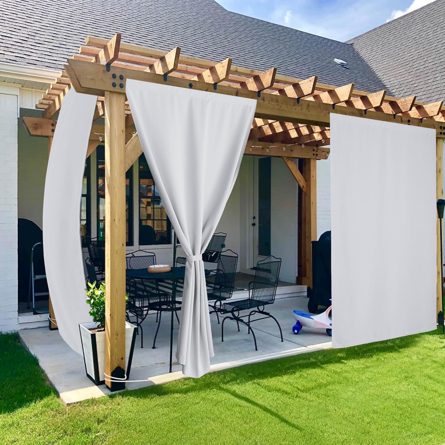 White privacy curtains draped on a wooden backyard pergola, with one tied back, offering shade to a patio dining area.