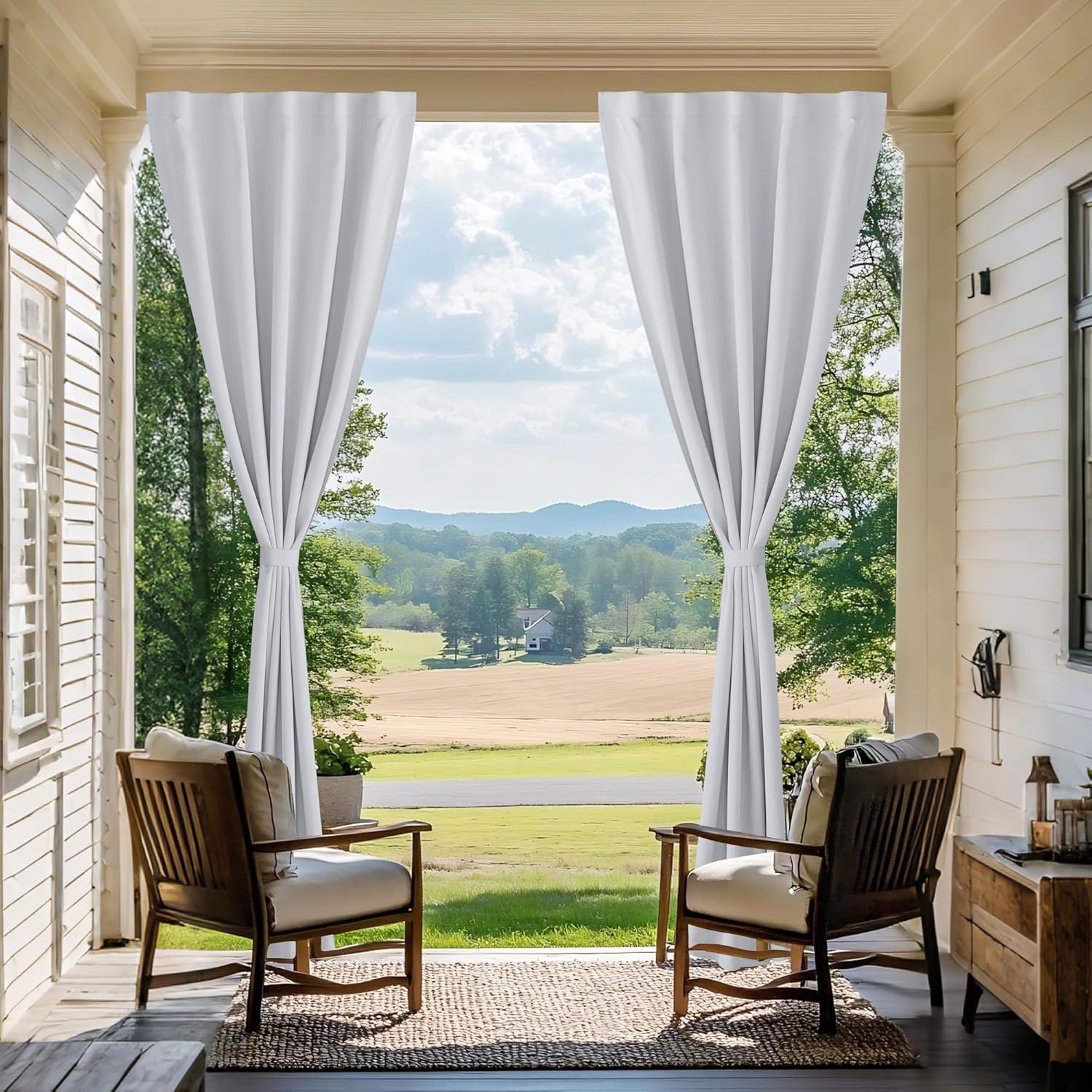 White outdoor curtains tied back on a porch, offering privacy and a framed view of green fields.