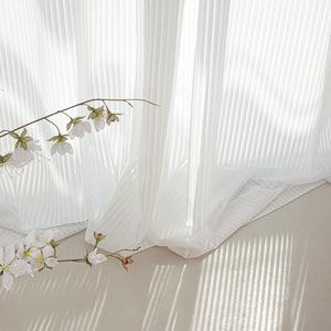 White pleated sheer curtains with natural light filtering through, beside a branch of white blossoms.