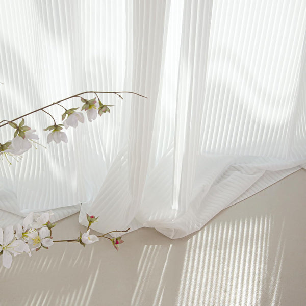 White pleated sheer curtains with natural light filtering through, beside a branch of white blossoms.