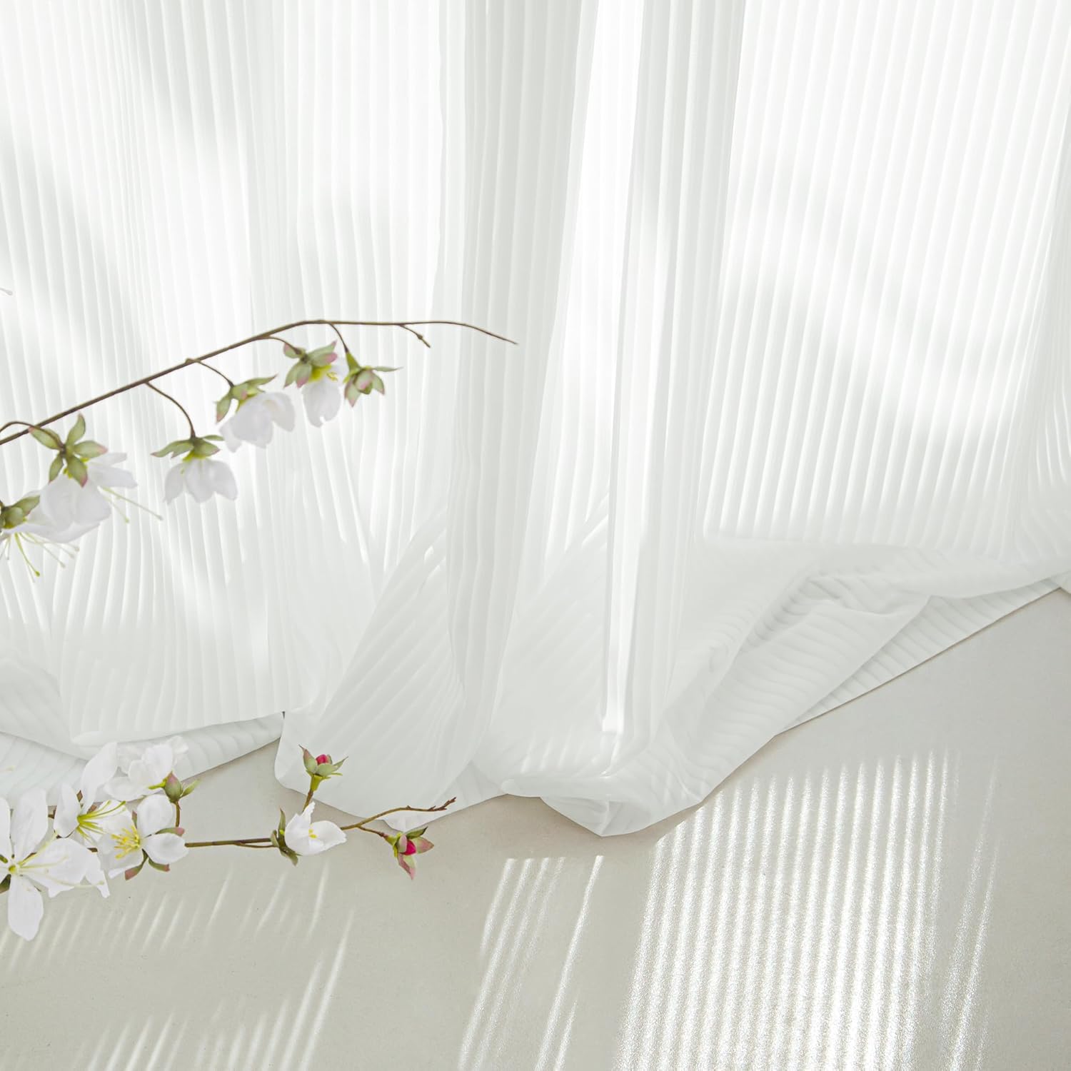 Elegant white pleated sheer curtains in natural light, with a branch of delicate white blossoms.