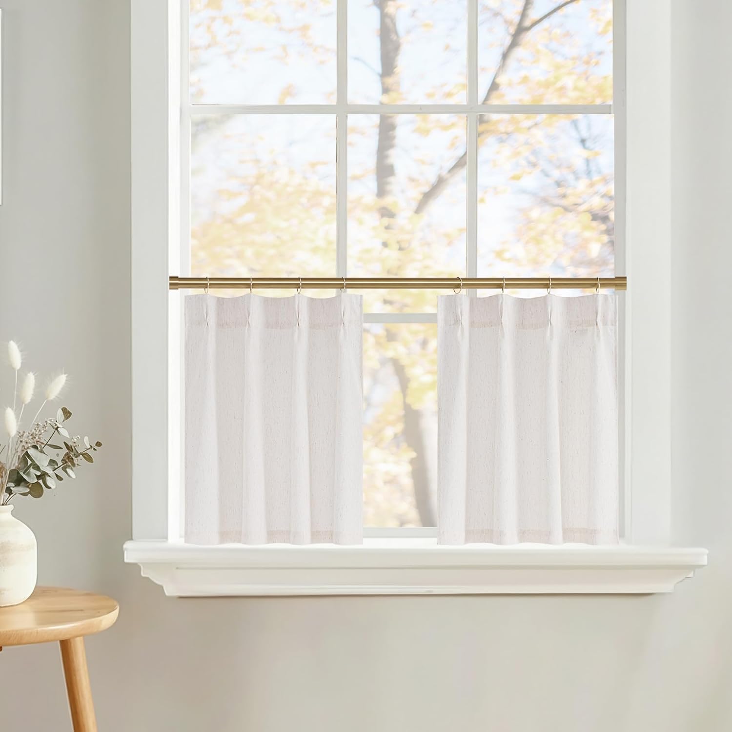 Sheer white pleated cafe curtains hanging from a gold rod in a bright room with a view of autumn trees.