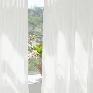 White vertically ribbed sheer curtains filtering soft daylight. Green trees and flowers visible outside.