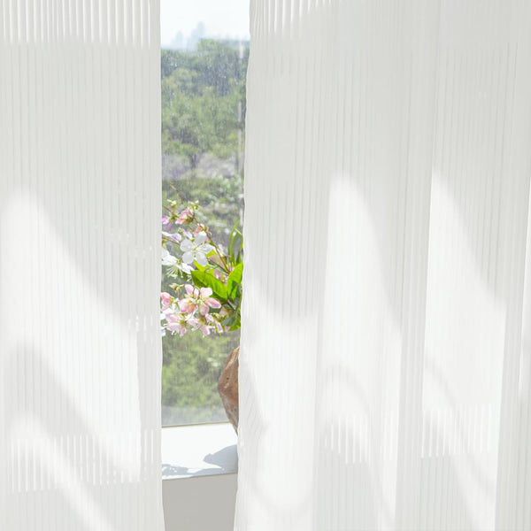 White vertically ribbed sheer curtains filtering soft daylight. Green trees and flowers visible outside.
